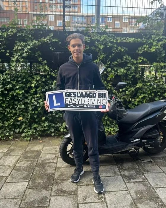 Geslaagde scooterleerling voor een groene heg, met het bord Geslaagd bij Easydriving voor een zwarte scooter