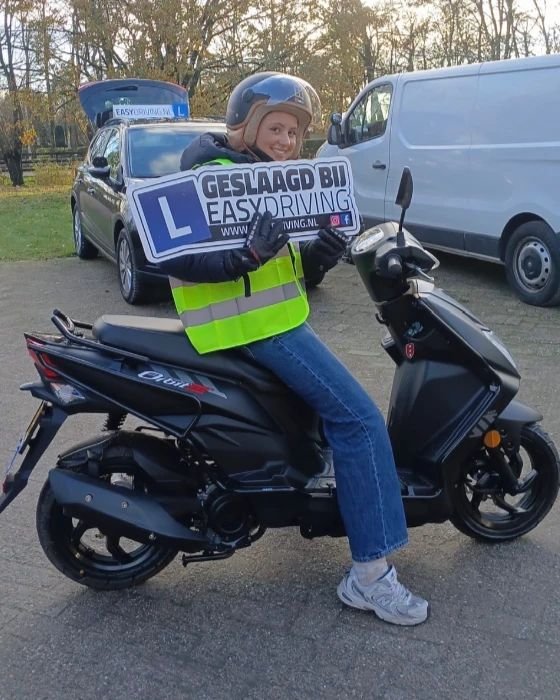 Geslaagde scooterleerling met helm op een zwarte scooter, met het bord Geslaagd bij Easydriving