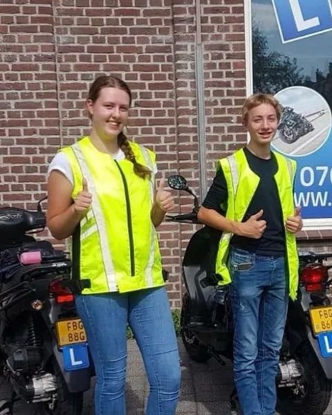 Twee geslaagde scooterleerlingen in gele hesjes steken hun duim op naast de zwarte lesscooters, voor het raam van de rijschool