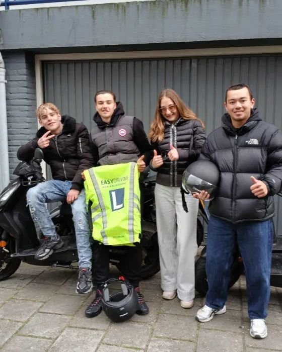 Vier scooterleerlingen voor een rij garages, een van hen houdt een hesje met het L van Easydriving omhoog