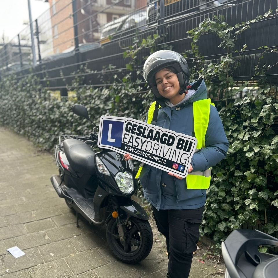 Geslaagde scooterleerling met helm en geel hesje houdt het bord Geslaagd bij Easydriving vast, naast een zwarte lesscooter